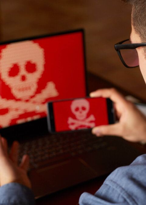 Man sits near laptop with phone blocked and encrypted by ransomware spyware asking for money. Laptop and smartphone infected by virus. Scary red skull crossbones on screen. Cyber security concept